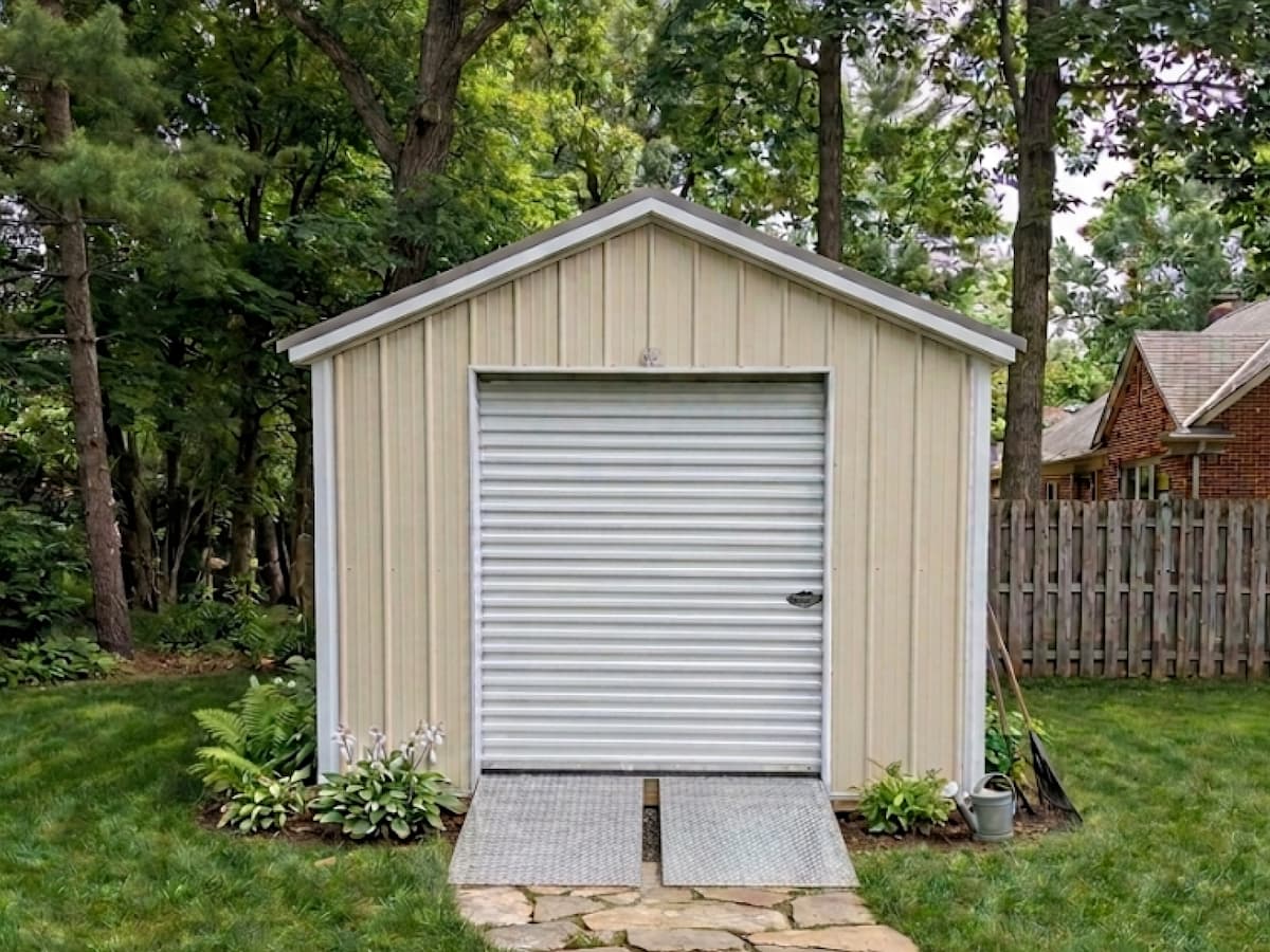 Tan Gable Shed with white trim and roll-up garage door in a wooded backyard