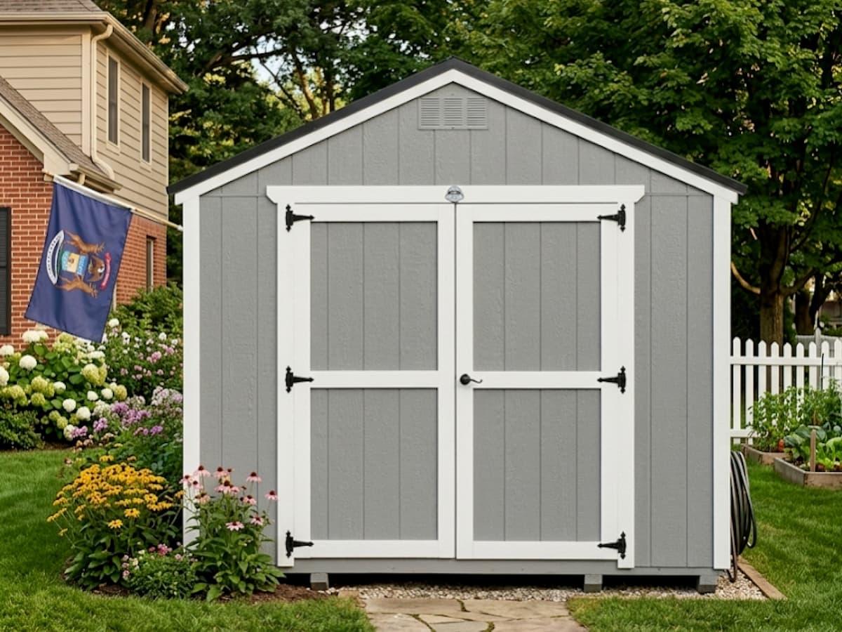 Gray Classic shed with white trim double doors beside a flower garden and Michigan flag